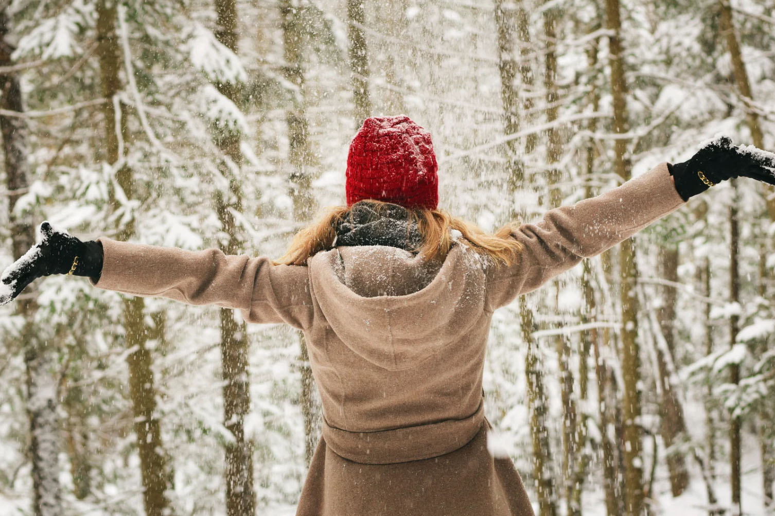 A person standing in a snowy forest, wearing a red hat and a beige coat, with arms outstretched, enjoying the snowfall.