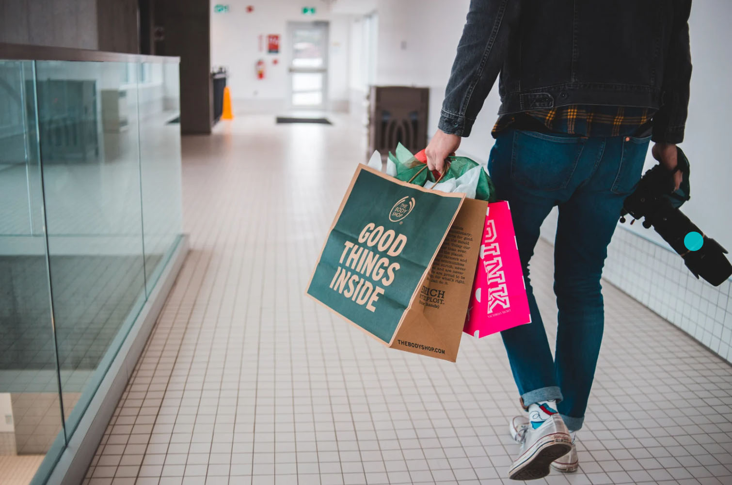 A person walking through a shopping mall carrying two shopping bags, one labeled "GOOD THINGS INSIDE" and the other from PINK, with a camera hanging from their neck.