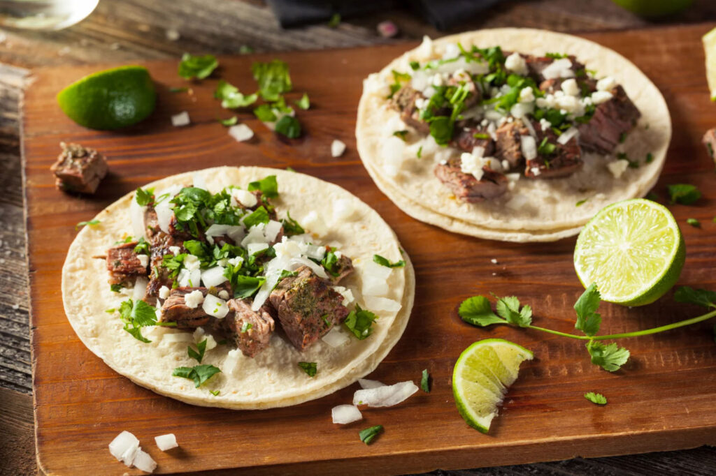 Two tacos filled with grilled beef, onions, and cilantro, garnished with lime wedges on a wooden surface.