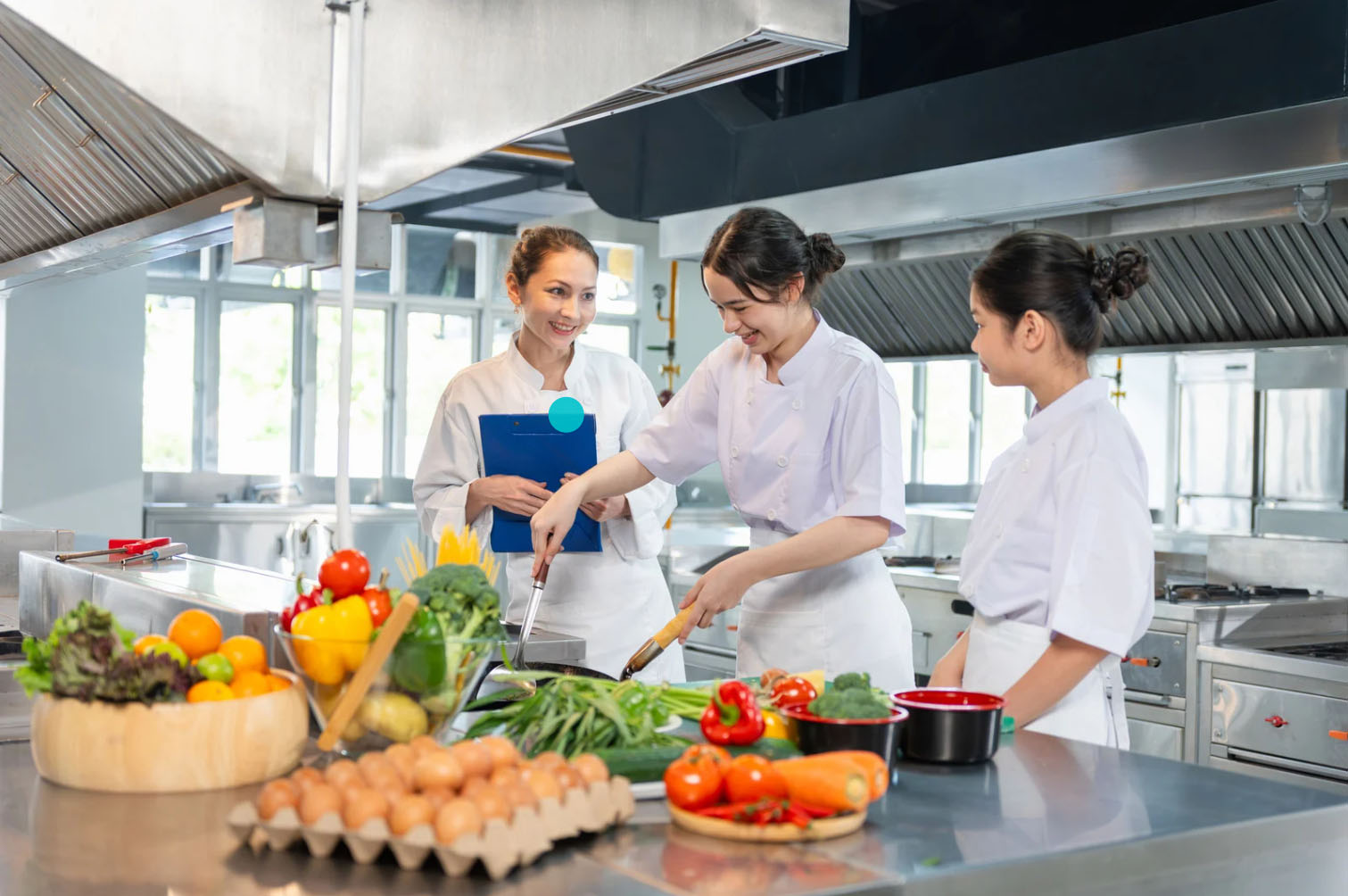 Three chefs in white uniforms working together in a professional kitchen, preparing fresh ingredients like vegetables, eggs, and fruits on the countertop.