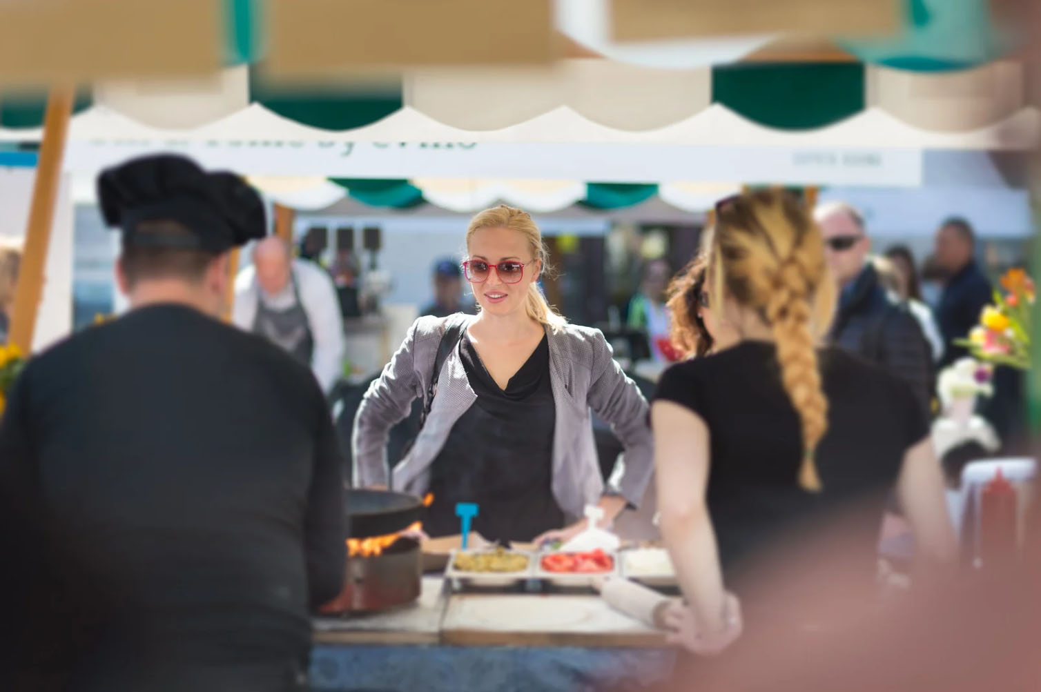 A woman with sunglasses stands at a food stall, engaging with customers while chefs prepare food at a lively outdoor market or food event.