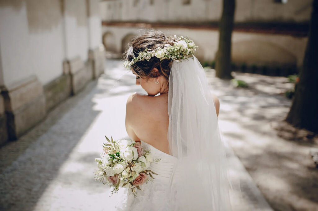 A bride walking down a sunny path, wearing a white wedding dress, a floral crown, and holding a bouquet of flowers.