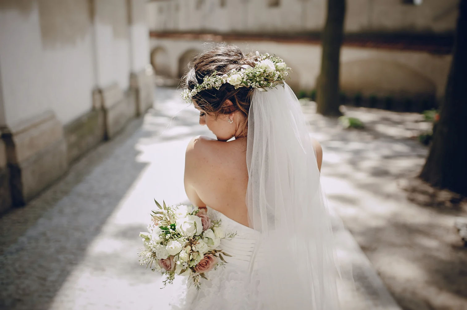 A bride walking down a sunny path, wearing a white wedding dress, a floral crown, and holding a bouquet of flowers.