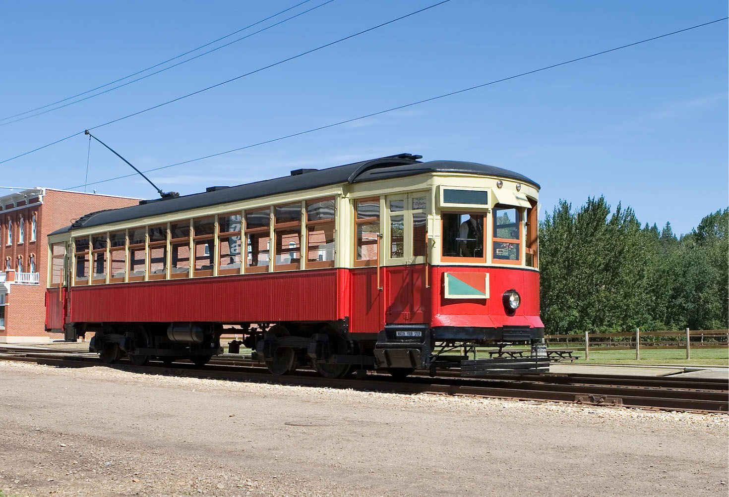 A vintage red and cream-colored streetcar parked on a railway track, set against a backdrop of a clear blue sky and green trees.