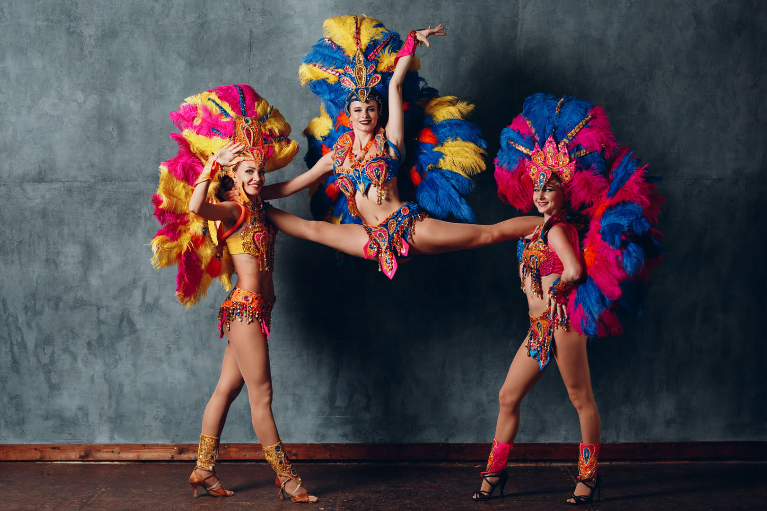 Three samba dancers in vibrant, colorful costumes with large feathered headdresses performing a lively dance at a carnival.