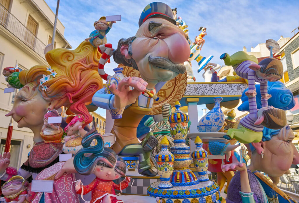 Colorful Las Fallas festival sculptures (ninots) in Valencia, Spain, depicting exaggerated cartoon-like figures and satirical characters arranged in a vibrant street display under a blue sky.