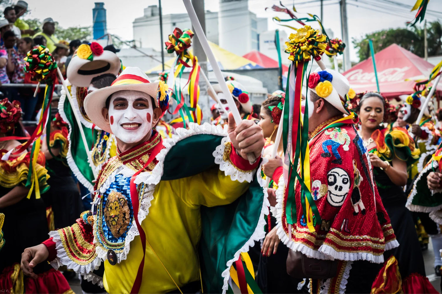 A joyful participant in the Barranquilla Carnival wearing colorful traditional attire and a painted face, leading the parade with other dancers in festive costumes.