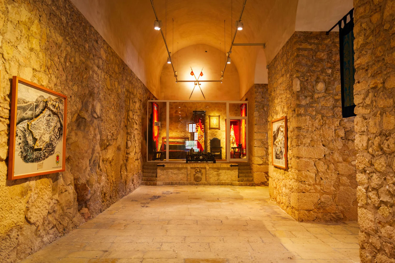 Stone-walled interior corridor of a historic museum or fortress, featuring framed maps and artifacts displayed along the walls under warm lighting, evoking medieval architecture and cultural heritage.