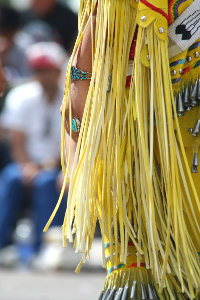 Close-up of a person wearing traditional Indigenous attire with yellow fringe and colorful beadwork, including a turquoise bracelet, with a blurred crowd in the background.