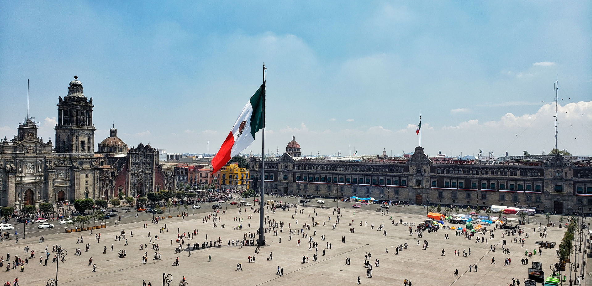 Wide daytime view of Mexico City’s Zócalo with the large Mexican flag flying in the center, historic cathedral and government buildings surrounding the busy public square.